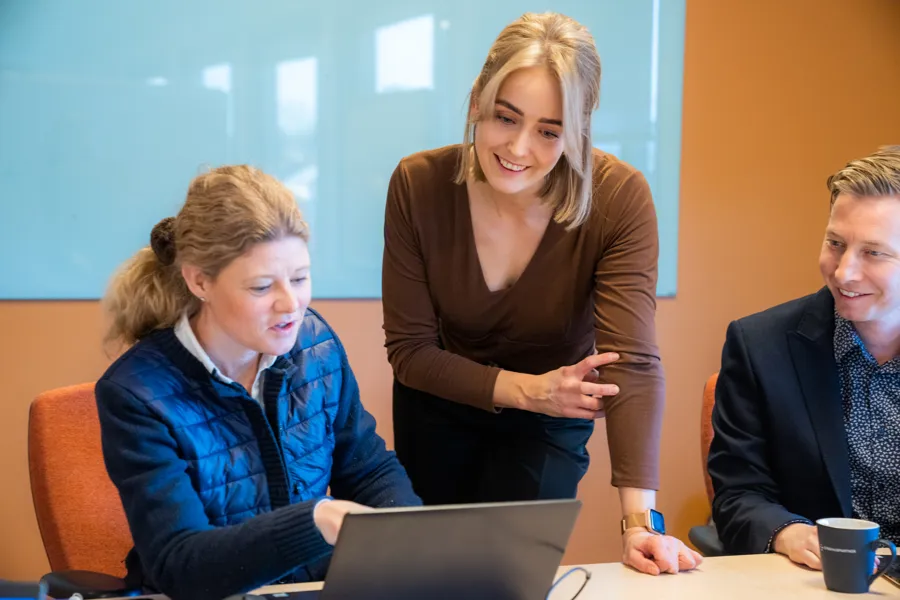 Tre personer foran en pc. En mann og en dame som sitter på hver sin side av en dame som står i midten. Foto.