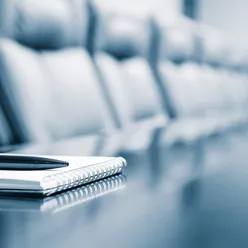 Skriveblokk på bord med mange stoler rundt. Foto.